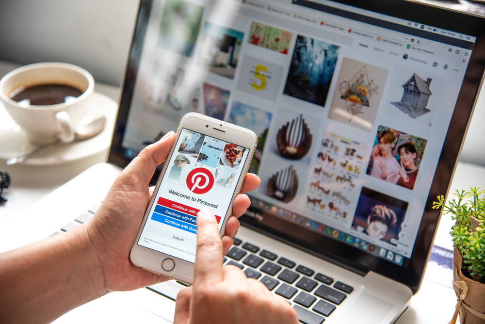 Pessoa acessando o Pinterest no smartphone enquanto trabalha em um laptop, com uma xícara de café ao lado e uma planta, ideal para buscar inspirações e ideias criativas.